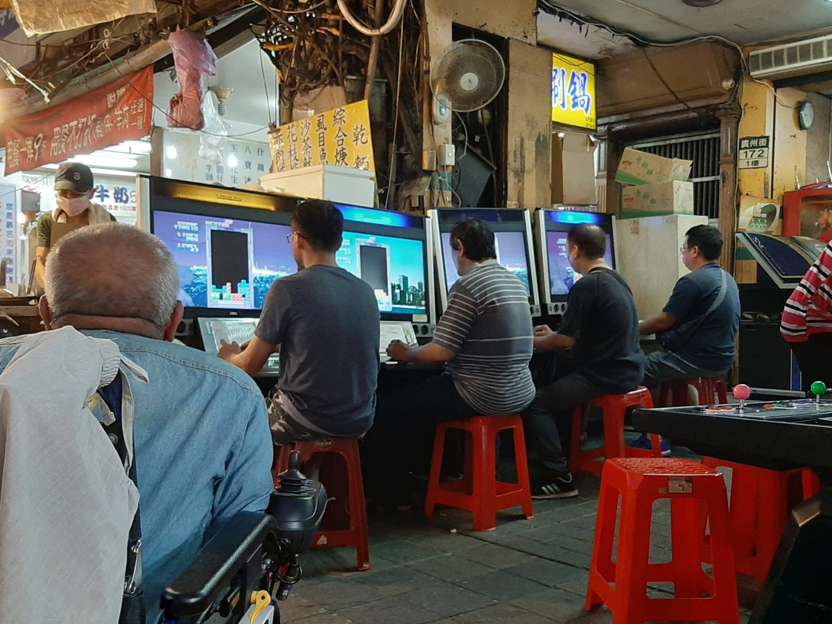 Several men at a Taiwanese night market playing Tetris on large computers.